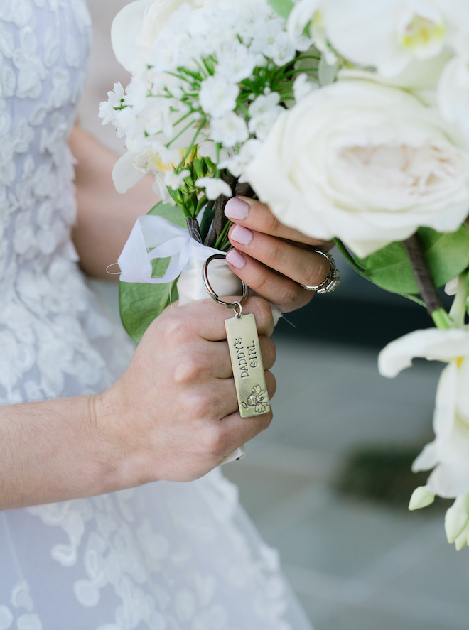 The bride holding her bouquet with white florals and a gold key chain that says "Daddy's Girl."