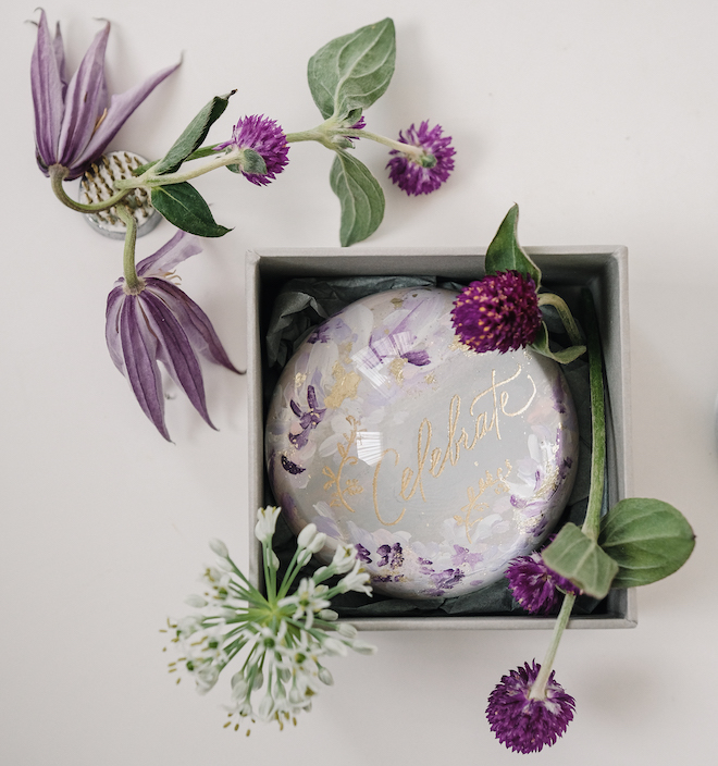 A clear paperweight with white and purple florals and "celebrate" written in gold in the middle. 