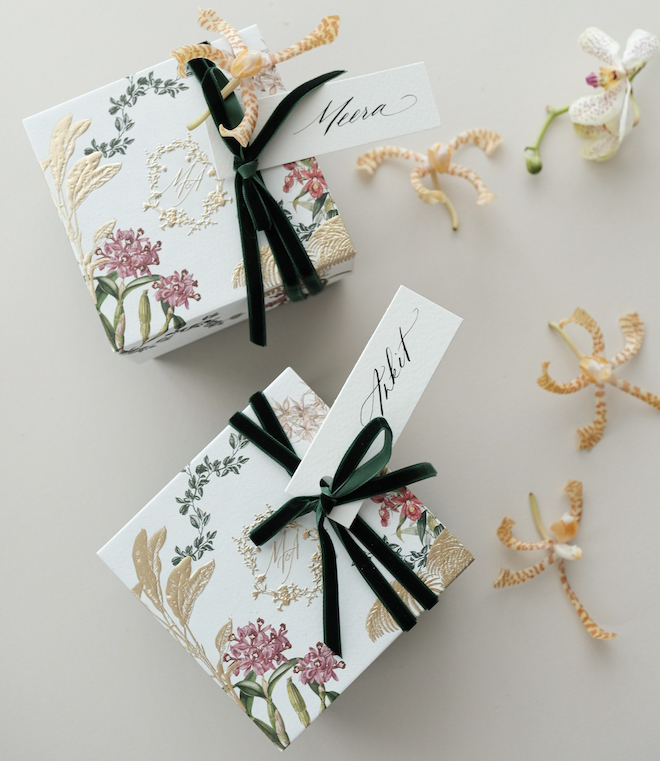 White boxes with gold, pink and green floral print on them. Black ribbon with name tags are tied on top.