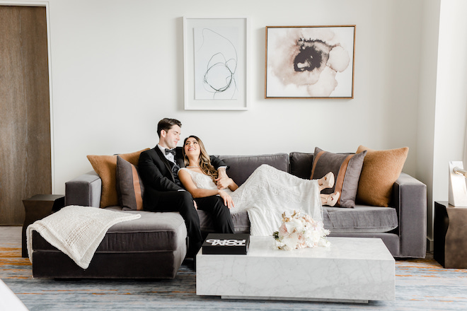 A bride and groom laughing and sitting on a gray couch in a living room.
