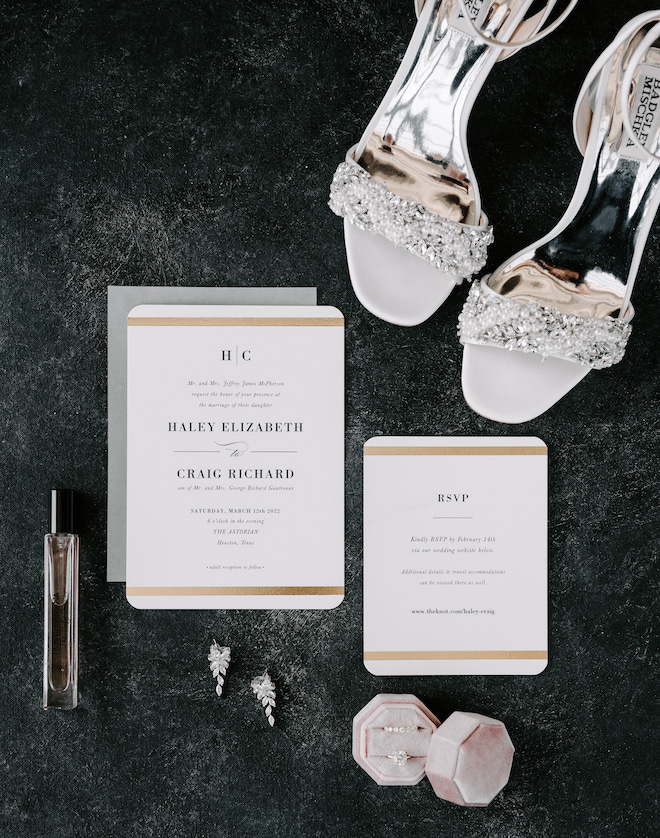 White invitations with gold detailing and black font. The brides shoes, perfume earrings and rings in a pink ring box sit next to it. 