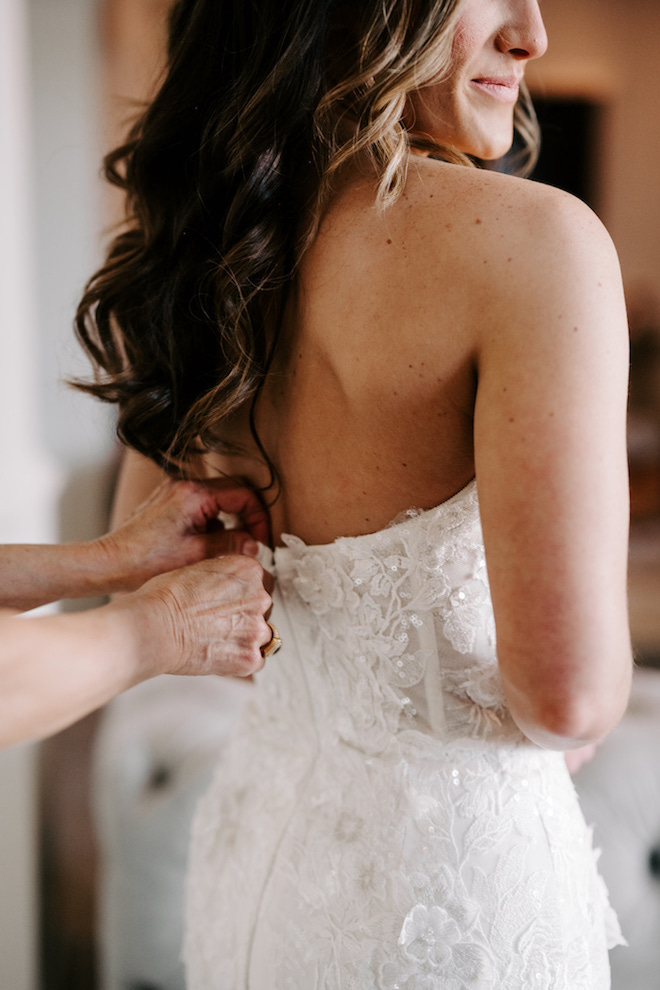 Hands zipping the back of the bride's strapless lace wedding gown. 