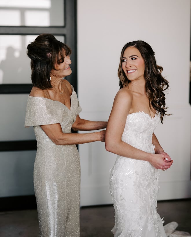 The brides mom buttoning up the back of her dress and the bride is smiling back at her. 