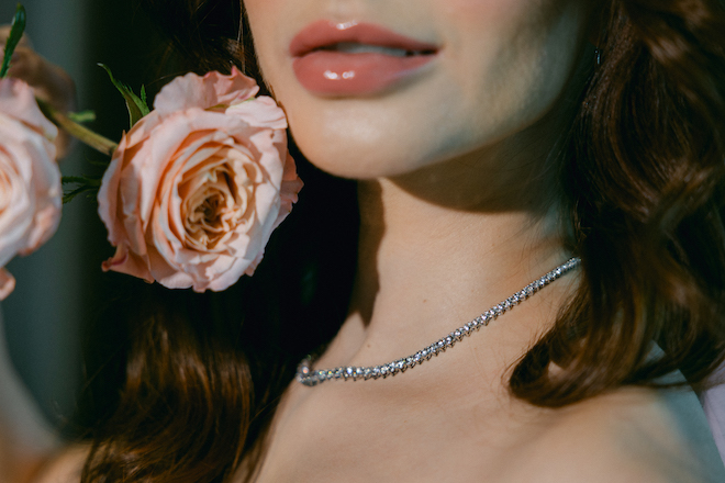 The brides diamond necklace resting on her chest while holding a pink flower up to her face. 