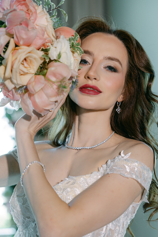 The bride holding a pink floral bouquet up to her face whole wearing red lipstick and a diamond necklace, earrings and bracelet.