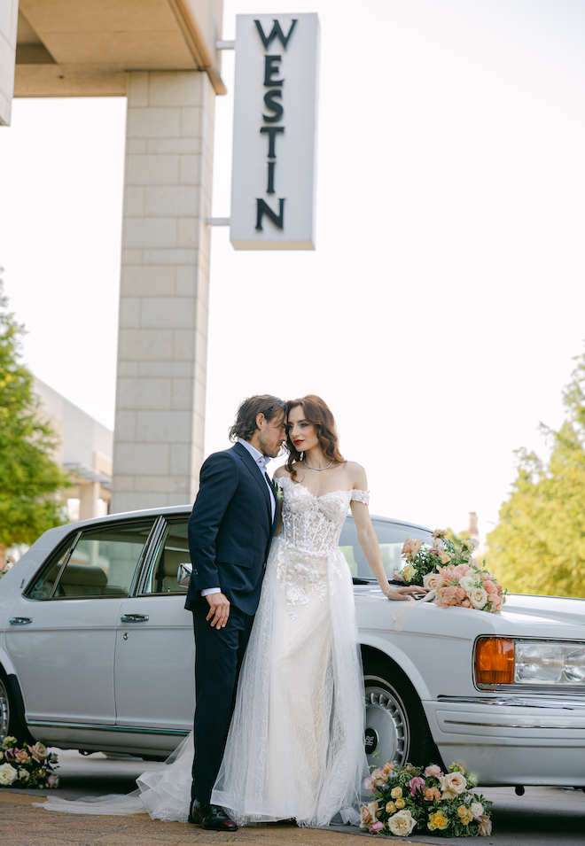 The bride and groom posing in front of a vintage Rolls Royce with pink and yellow floral arrangements decorating it in front of The Westin Houston Memorial City sign. 