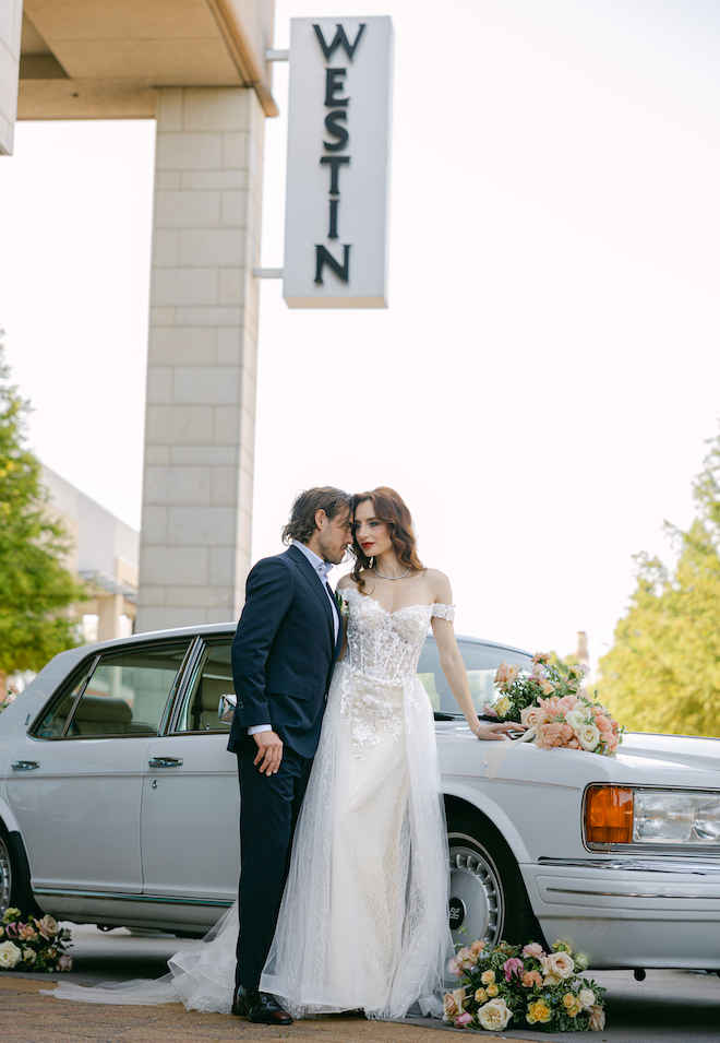 The bride and groom posing in front of a vintage Rolls Royce with pink and yellow floral arrangements decorating it in front of The Westin Houston Memorial City sign.