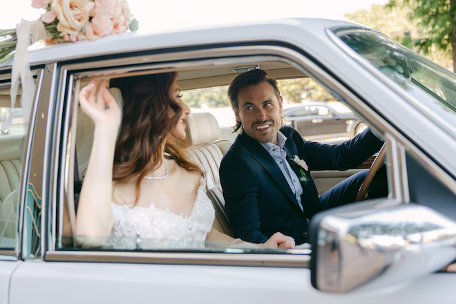 The groom sitting in the driver's seat of the vintage Rolls Royce smiling at the bride in the passenger seat. 