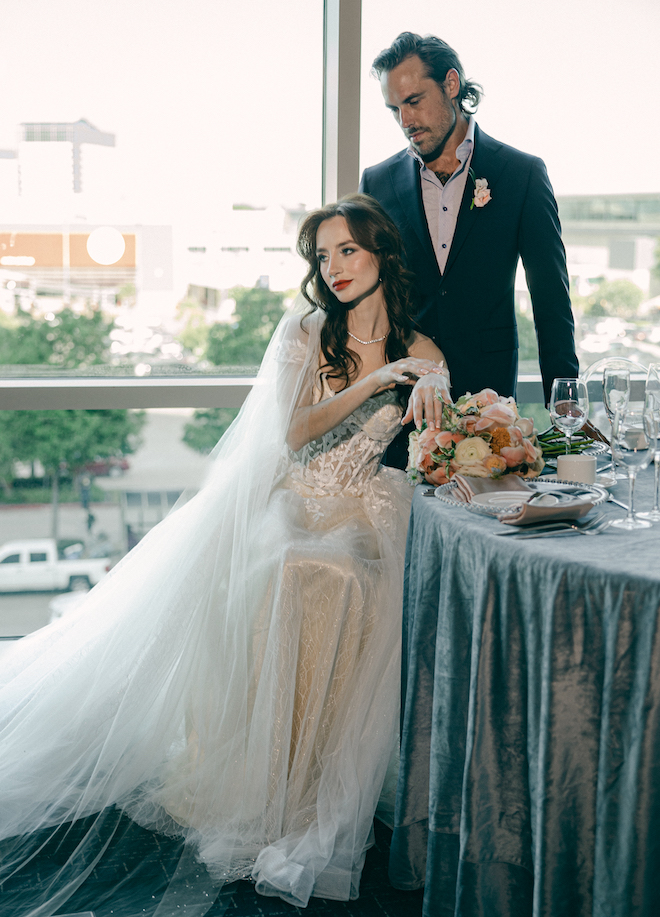 The bride sitting at the reception table with the groom standing behind looking down at her with floor-to-ceiling windows behind them in the ballroom of The Westin Houston Memorial City.