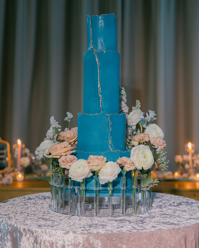 A four-tier blue cake with gold trim sitting in an acrylic cake stand decorated with pink and white florals. 