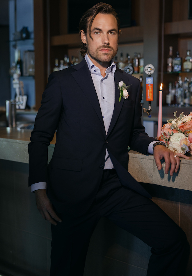 The groom standing at the bar in The Westin Houston Memorial City wearing a blue suit from b.Kreps&Co.