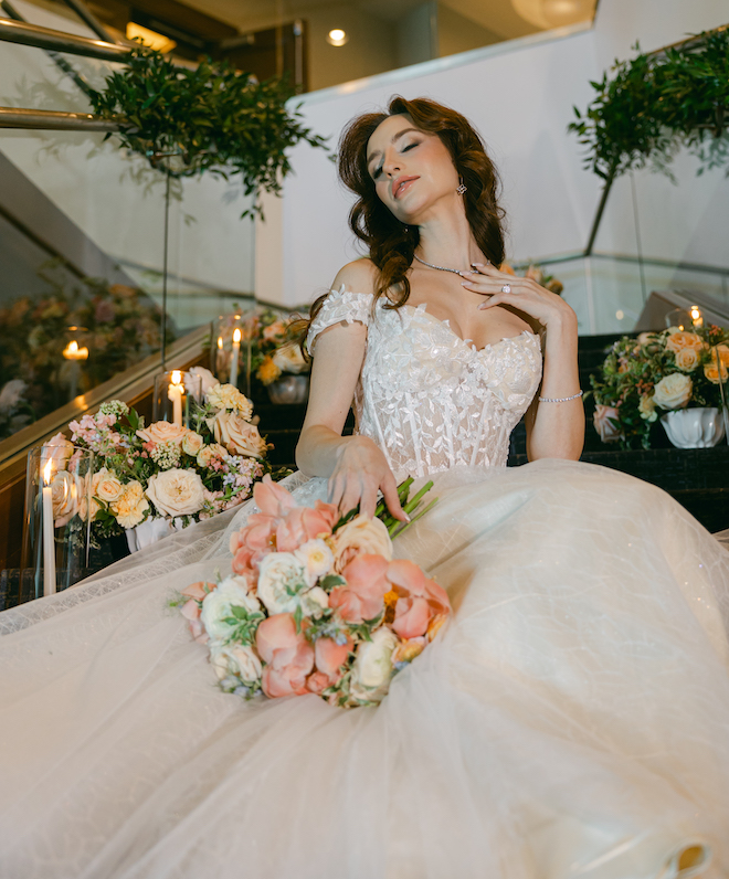 The bride touching her diamond necklace holding a pink and white bouquet. She is sitting on a staircase covered on florals and candles.