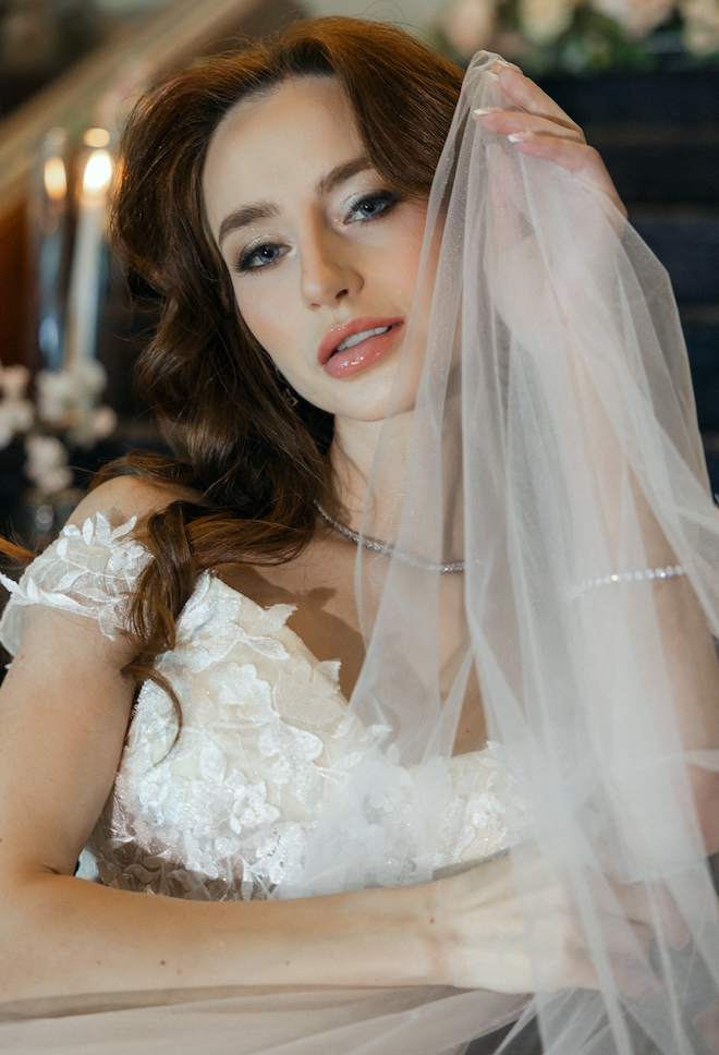 The bride wearing a floral applique wedding dress holding her veil up to her face. 