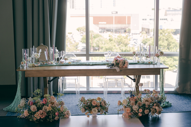 A wooden rectangle reception table decorated with pink and green floral arrangements and pink and white candles in front of the floor-to-ceiling windows in the ballroom of The Westin Houston Memorial City.