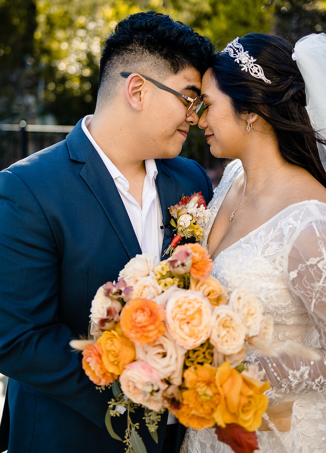 The bride and groom smiling and pressing their foreheads together. The bride is holding a bouquet with orange, sand and peach florals. 