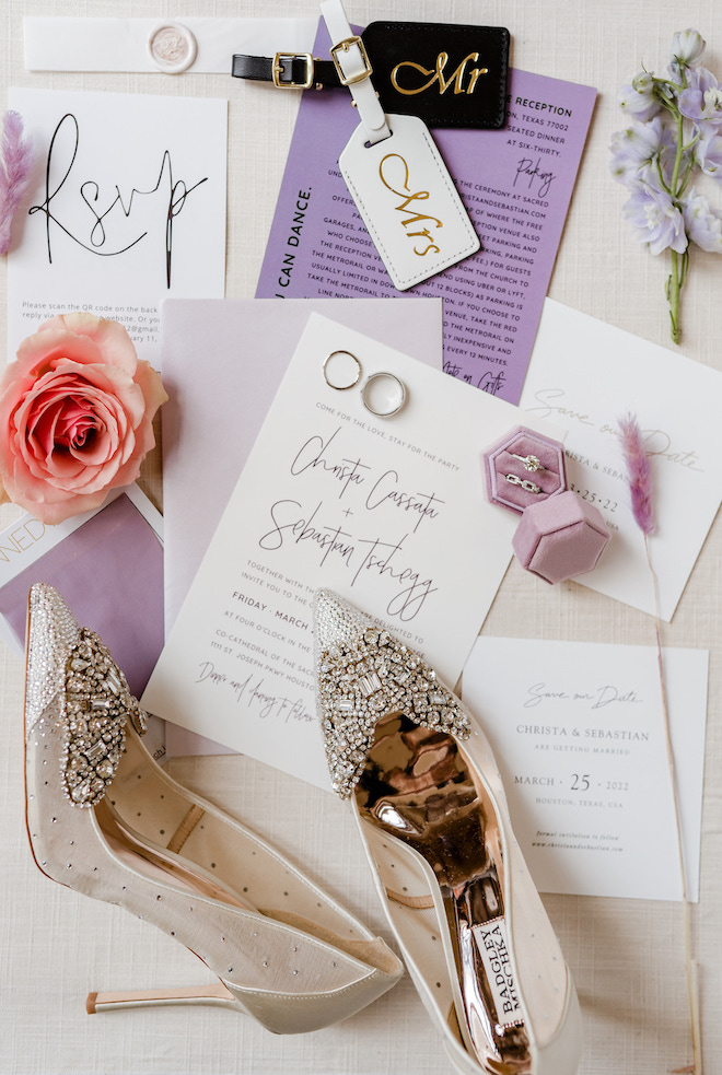 A white and lavender invitation suite with gold shoes, a pink ring box, pink and purple flowers and Mr. and Mrs. luggage tags.