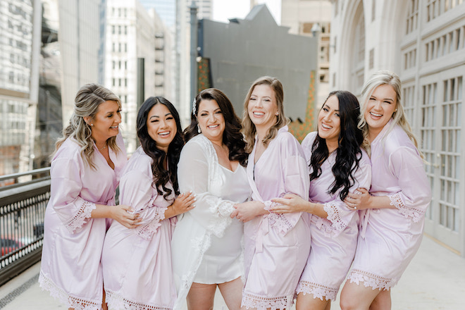 The bride wearing a white silk robe hugging her five bridesmaids wearing lavender silk robes. 