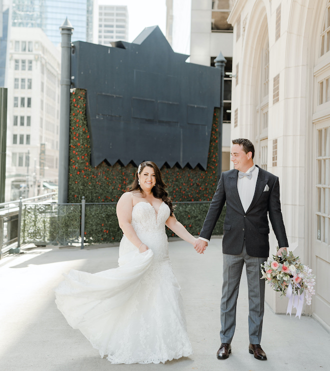 A bride and groom holding hands and the groom holding a bouquet in his other hand, standing on the terrace of the historic Crystal Ballroom at the Rice.