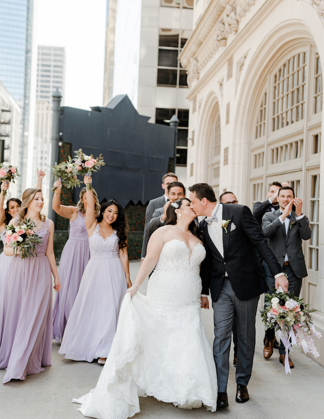 The bride and groom kissing with the wedding party in lavender dresses and gray suits cheering and clapping behind them.
