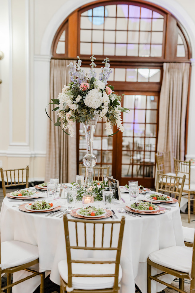 A round reception table with a white tablecloth and a purple, pink and white floral centerpiece. 