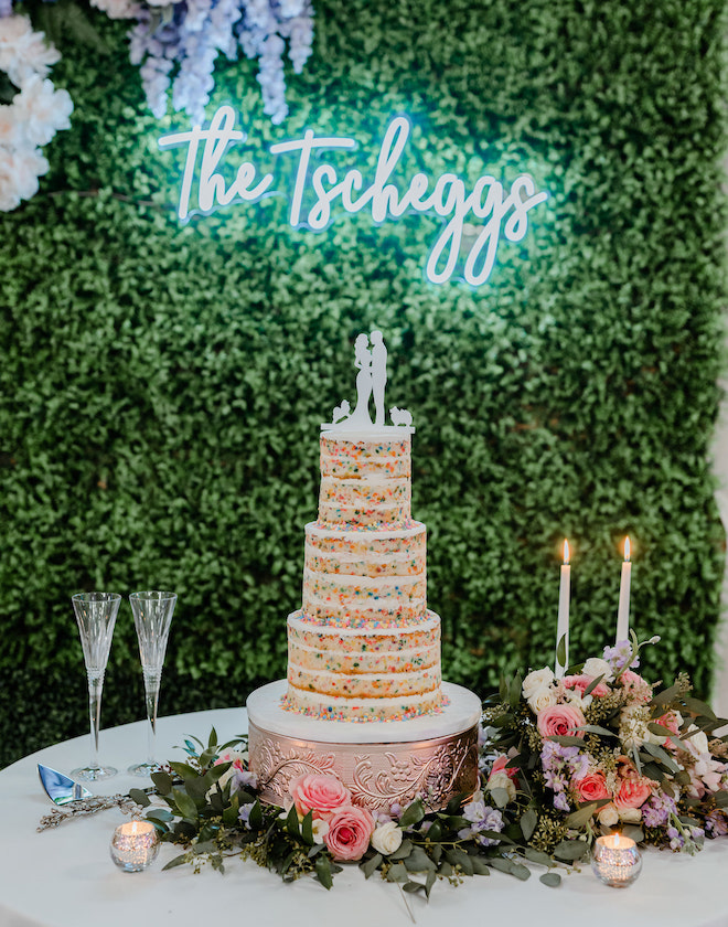 A three-tier confetti cake by Cakes by Gina in front of a greenery wall with "The Tscheggs" in lit-up marquee letters.