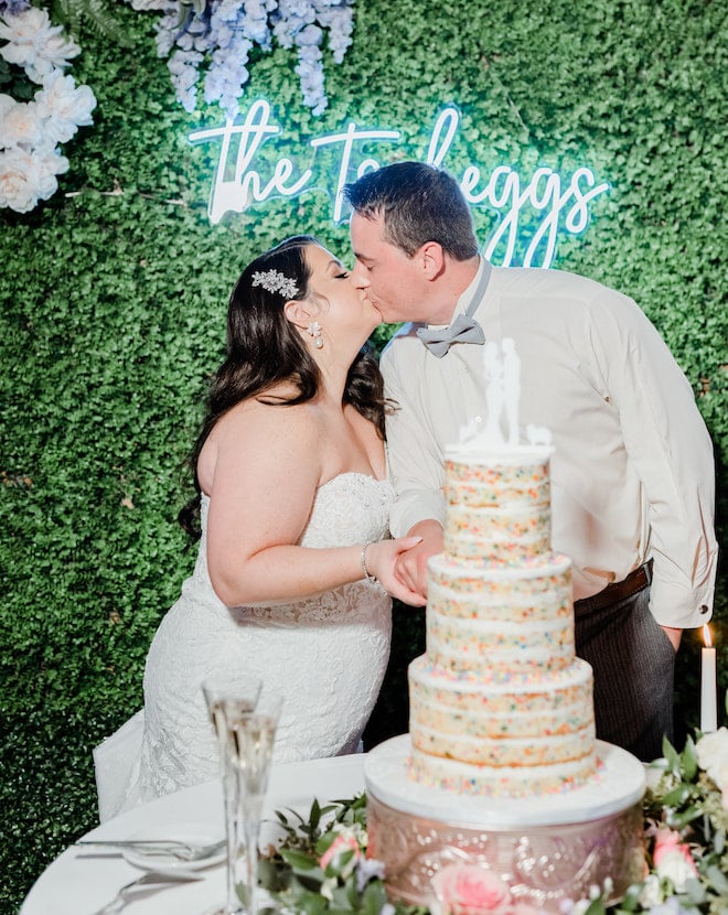 The bride and groom kissing while cutting a three-tier confetti cake. 