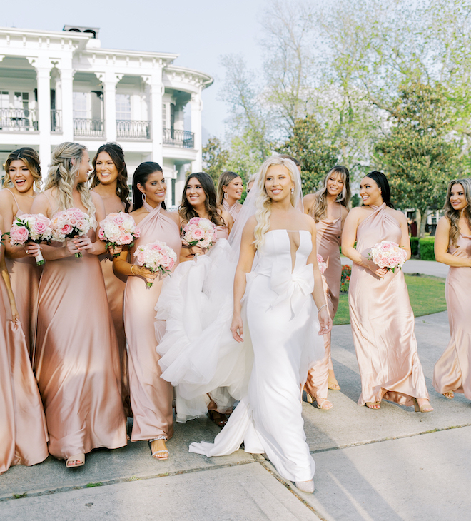 A bride walking with her bridesmaids in pink satin dresses and white and pink bouquets.