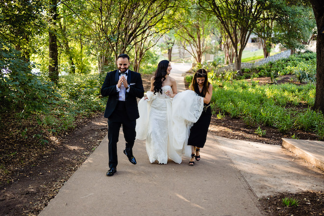 Wedding planner Marcela Bogado holding the bride's wedding gown as they walk with the groom on a shaded sidewalk. 