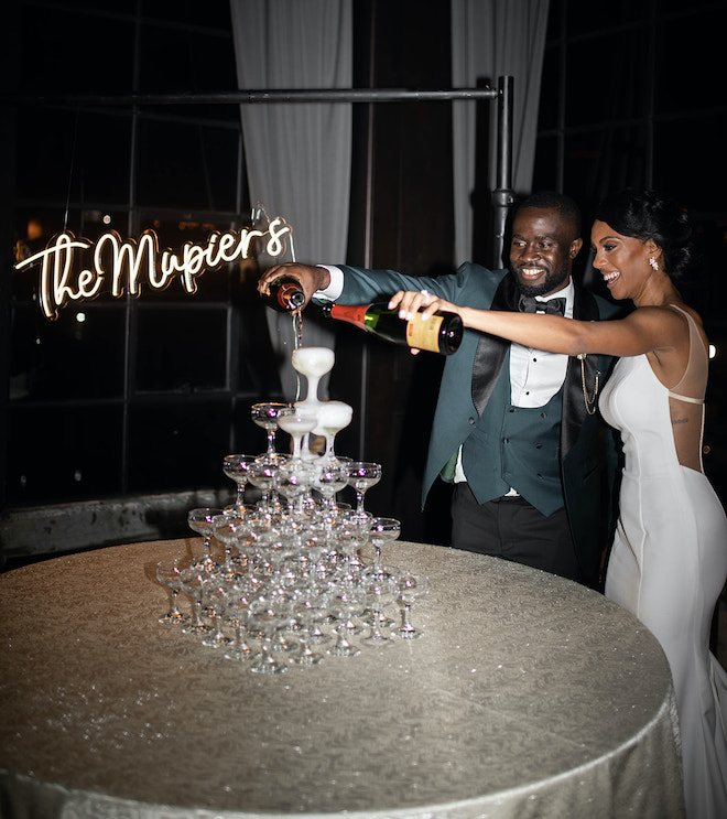 The bride and groom pouring champagne into a tower of glasses with a lit-up sign that says "The Mupier's."