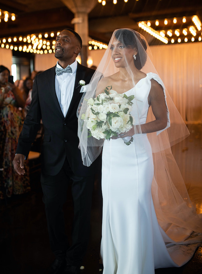 The bride wearing a veil over her head and holding a white and green bouquet walking down the aisle with her father. 