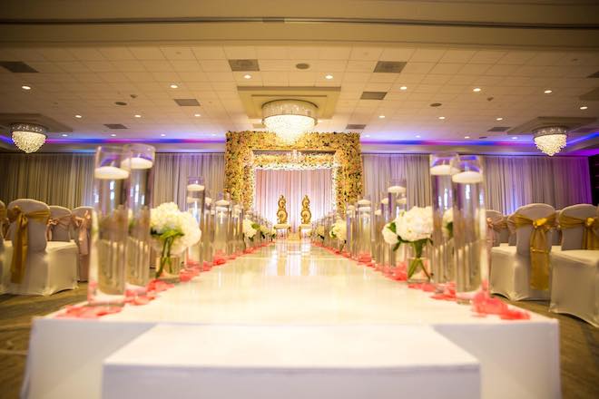 A white aisle leading to a floral Mandap in a ballroom.