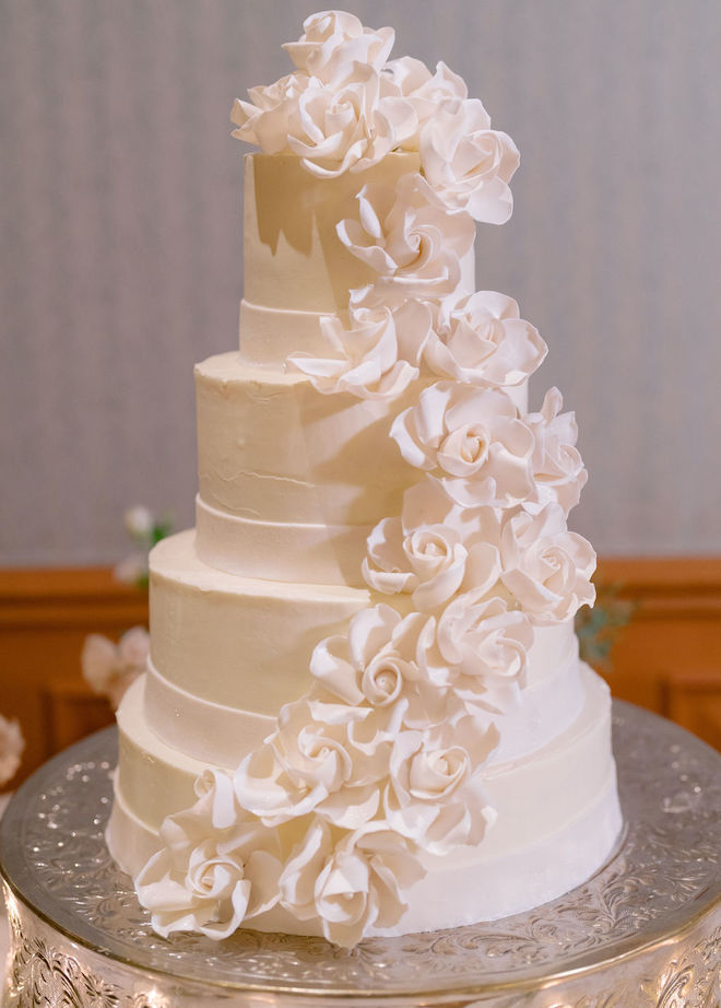 A 4-tier wedding cake detailed in white roses rests on a silver cake stand at the wedding reception. 