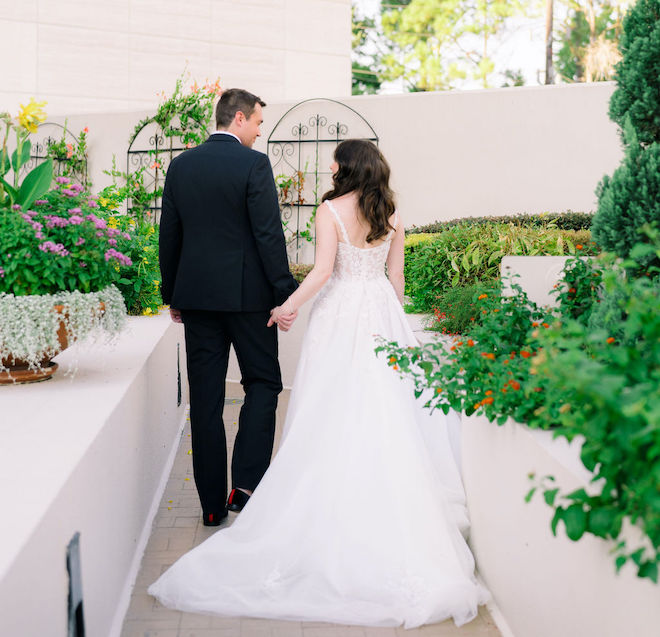 The bride and groom hold hands as they walk through the hotel's outdoor space, thriving with flowers and greenery.