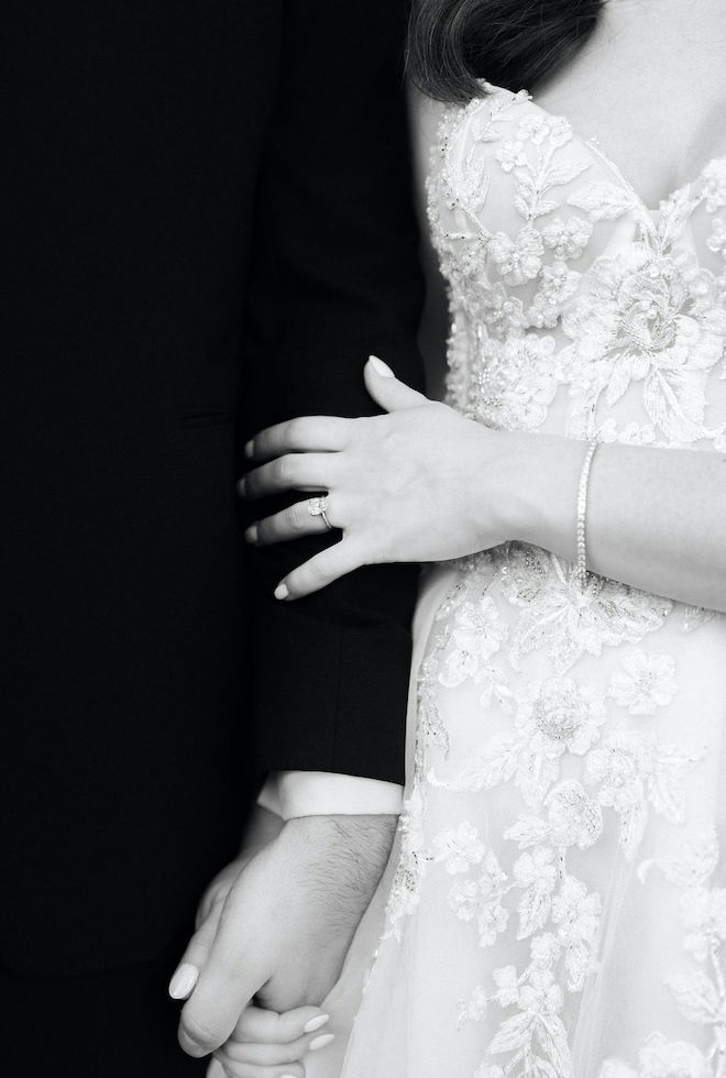 A close-up, black and white photo of the bride and groom holding hands on their wedding day. 