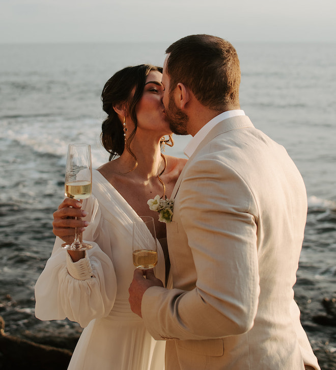 The bride and groom kissing while holding champagne glasses overlooking the ocean.