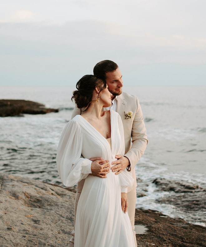 The bride and groom standing on a cliff looking at the ocean after their sunset elopement. 