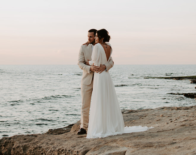 A bride and groom hugging on a cliff overlooking the ocean after their sunset elopement. 