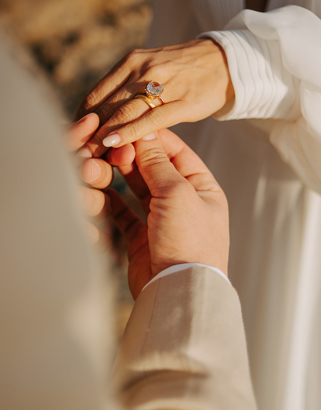 The groom putting on a gold wedding band and diamond engagement ring on the bride's hand. 