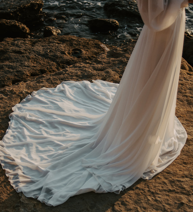 The bottom of the brides white wedding gown while standing over a cliff overlooking the ocean. 