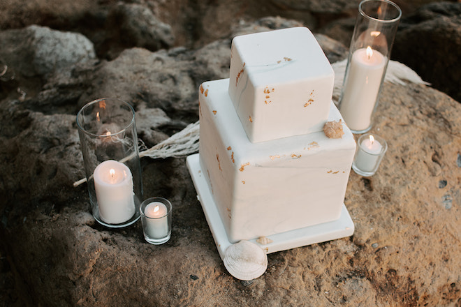 A two-tier white cake with gold specs surrounded by candles for an intimate elopement on the beach. 