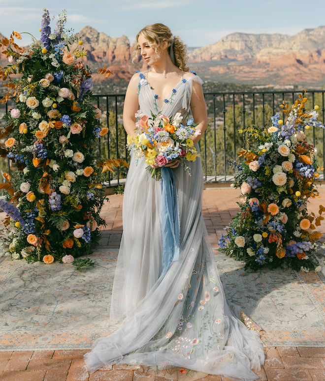 The bride wearing a blue dress and holding a bouquet of blue and citrus-colored florals. She is standing in front of the altar overlooking the Sedona mountains. 