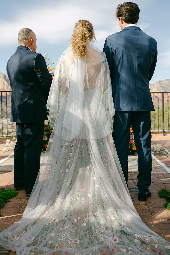 The bride, groom and officiant facing the mountains showing the back of the bride's blue gown with florals on the train. 