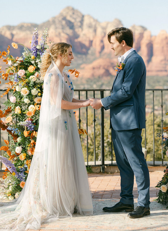 The bride and groom holding hands during their ceremony with vibrant citrus and blue florals behind them and a mountain view. 
