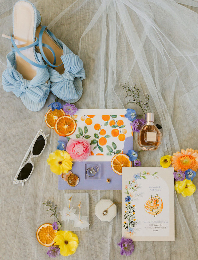 A citrus-themed invitation suite with blue shoes, fresh orange slices, bridal jewelry and white sunglasses.