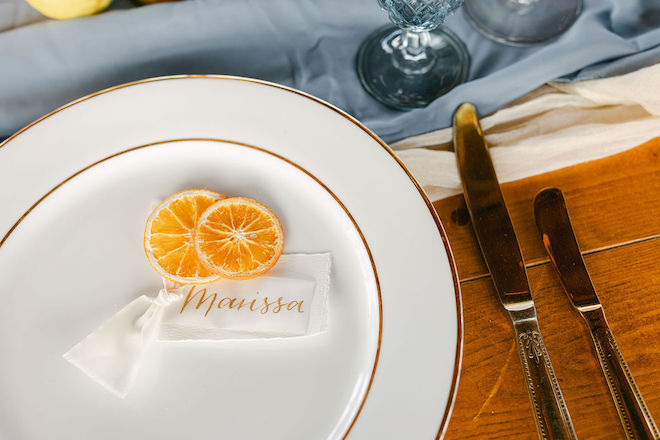 A white plate with gold lining with flatware. Two orange slices and a name card reading "Marissa" sit on the plate. 