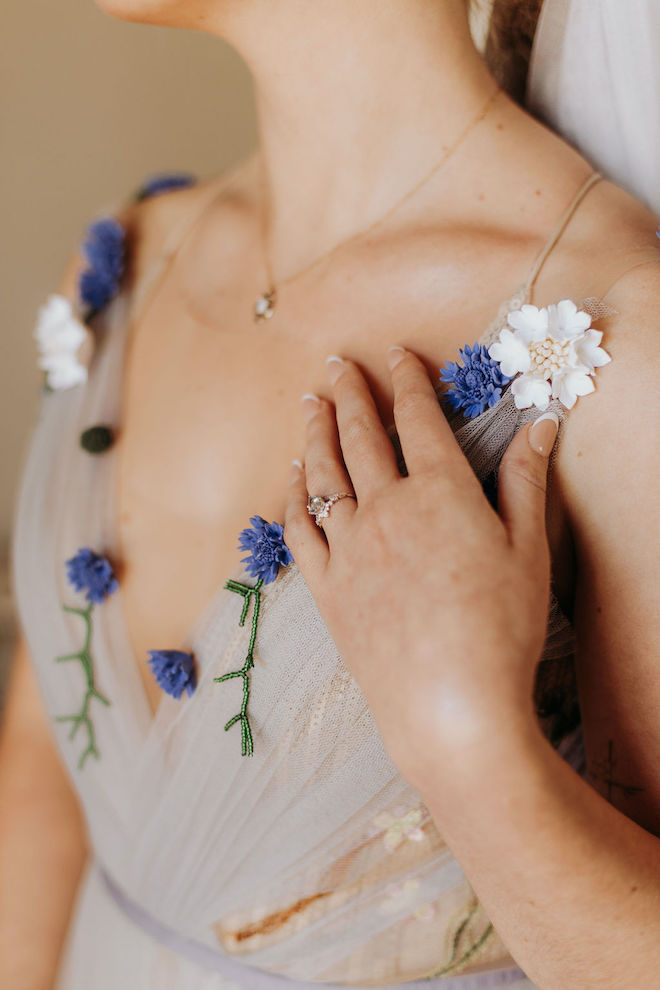 The bride holding her hand up to her blue floral wedding gown, showing her engagement ring. 