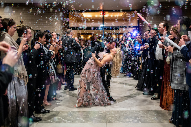 The bride and groom kissing during a bubble send off. The bride changed into a pink floral dress. Guests are cheering on either side of them.