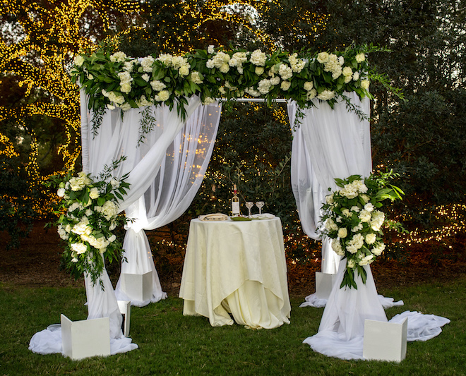 A white Chuppah with white and green floral arrangements in front of a lit up oak tree. 