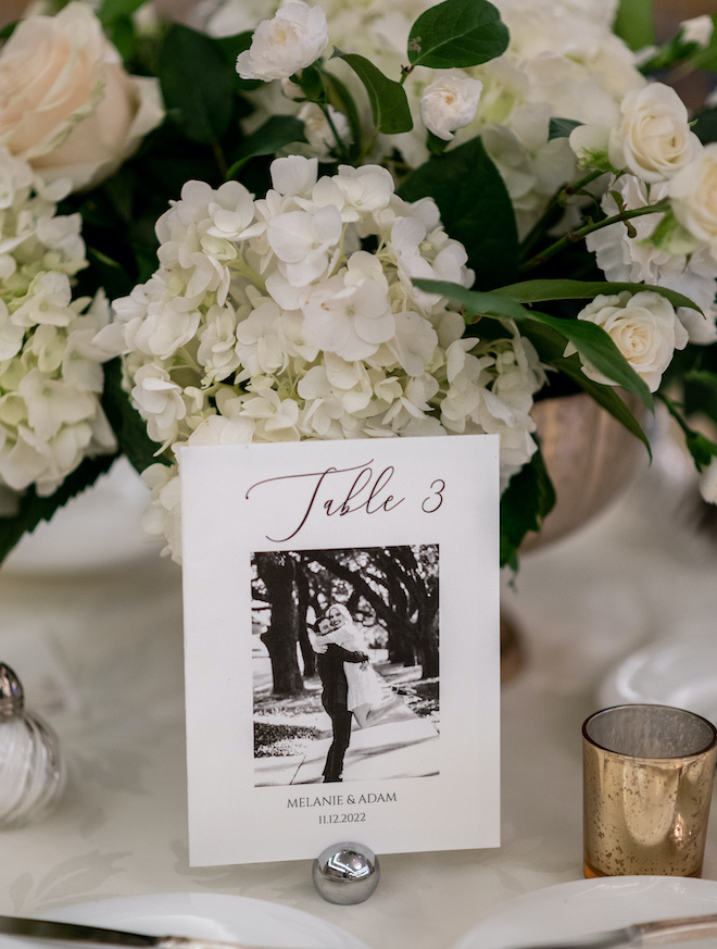 A photo of the bride and groom and the table number in front of a white and green floral arrangement. 