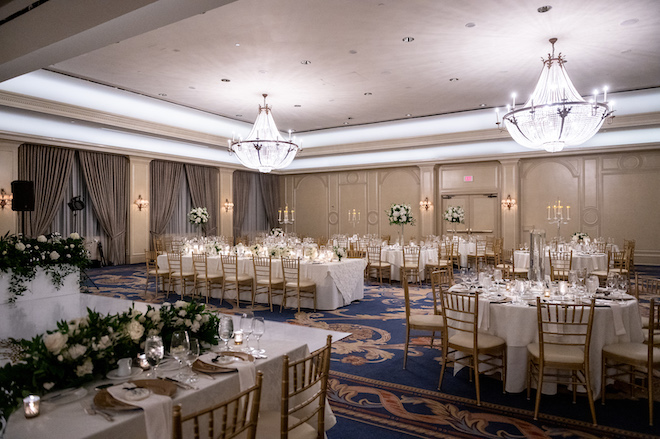 The ballroom reception with white tables and gold chairs and green and white floral arrangements.